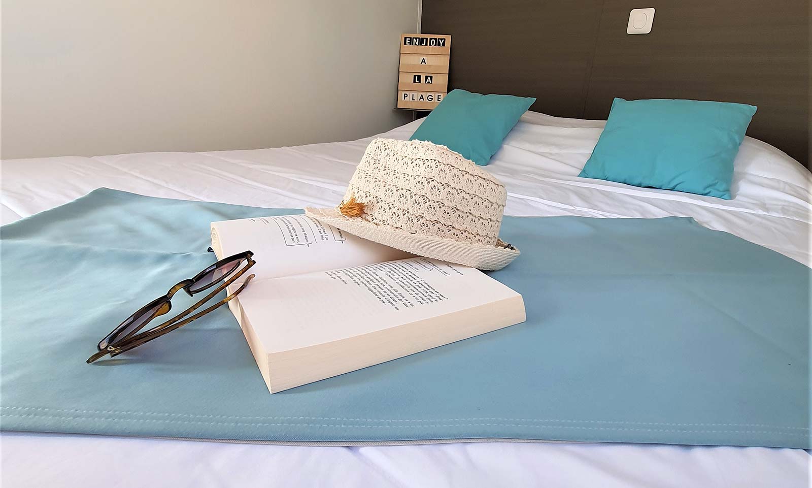 Livre, chapeau de paille et lunettes de soleil sur lit double d'un mobil-home à Saint-Hilaire
